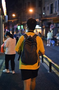 A man with a backpack walks through the lively streets of Xiamen at night.