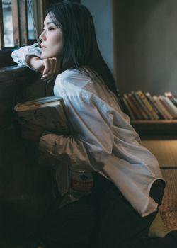 Asian woman in white shirt holds a book, gazing wistfully by a window indoors.