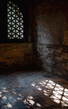 Sunlight casts intricate patterns through a historic window grille onto a brick floor, creating a serene atmosphere.