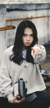 Young woman pointing while holding a vintage camera outdoors with a serious expression.