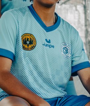 A young soccer player wearing a light blue team jersey with club logos, sitting outdoors.