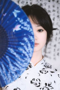 Half face portrait with blue fan, calligraphy background, traditional attire.