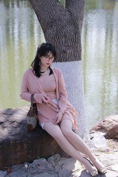 A stylish woman in pink sits by a tree near Xuanwu Lake, Nanjing, enjoying a sunny day.