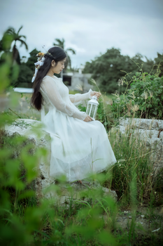 Woman in a flowing white dress holding a lantern, seated in a lush outdoor setting.