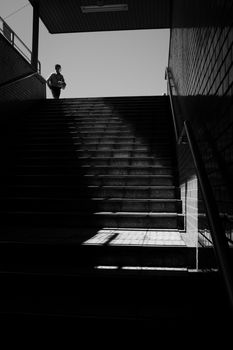 Moody black and white silhouette of a man standing at the top of shadowed stairs outdoors.