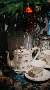 A vintage porcelain teapot with cups and Christmas ornaments under a decorated tree branch.