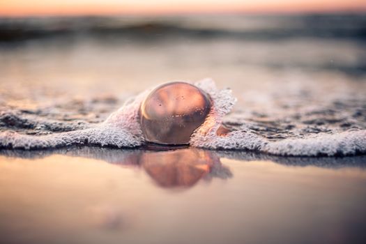A glass ball reflects the ocean sunset as waves crash around it on a beach in Świnoujście.